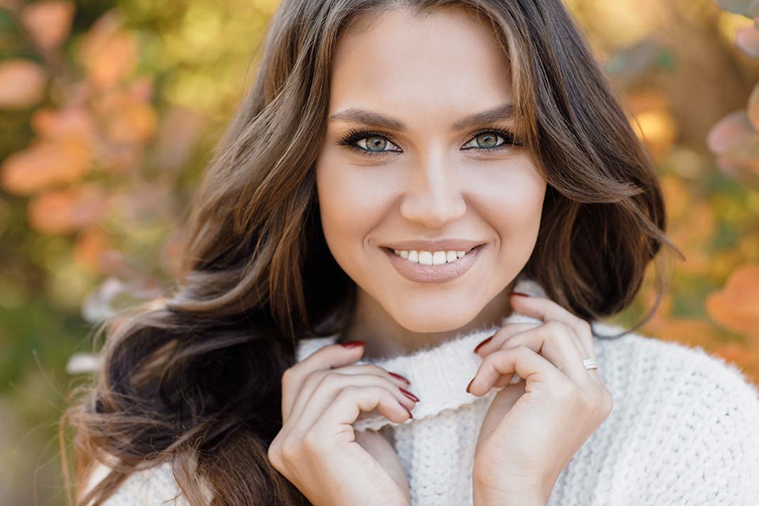 Smiling woman with long, wavy brown hair and bright eyes, wearing a cozy sweater in an outdoor setting, representing the natural and radiant results of treatments at Revive Aesthetics Med Spa.